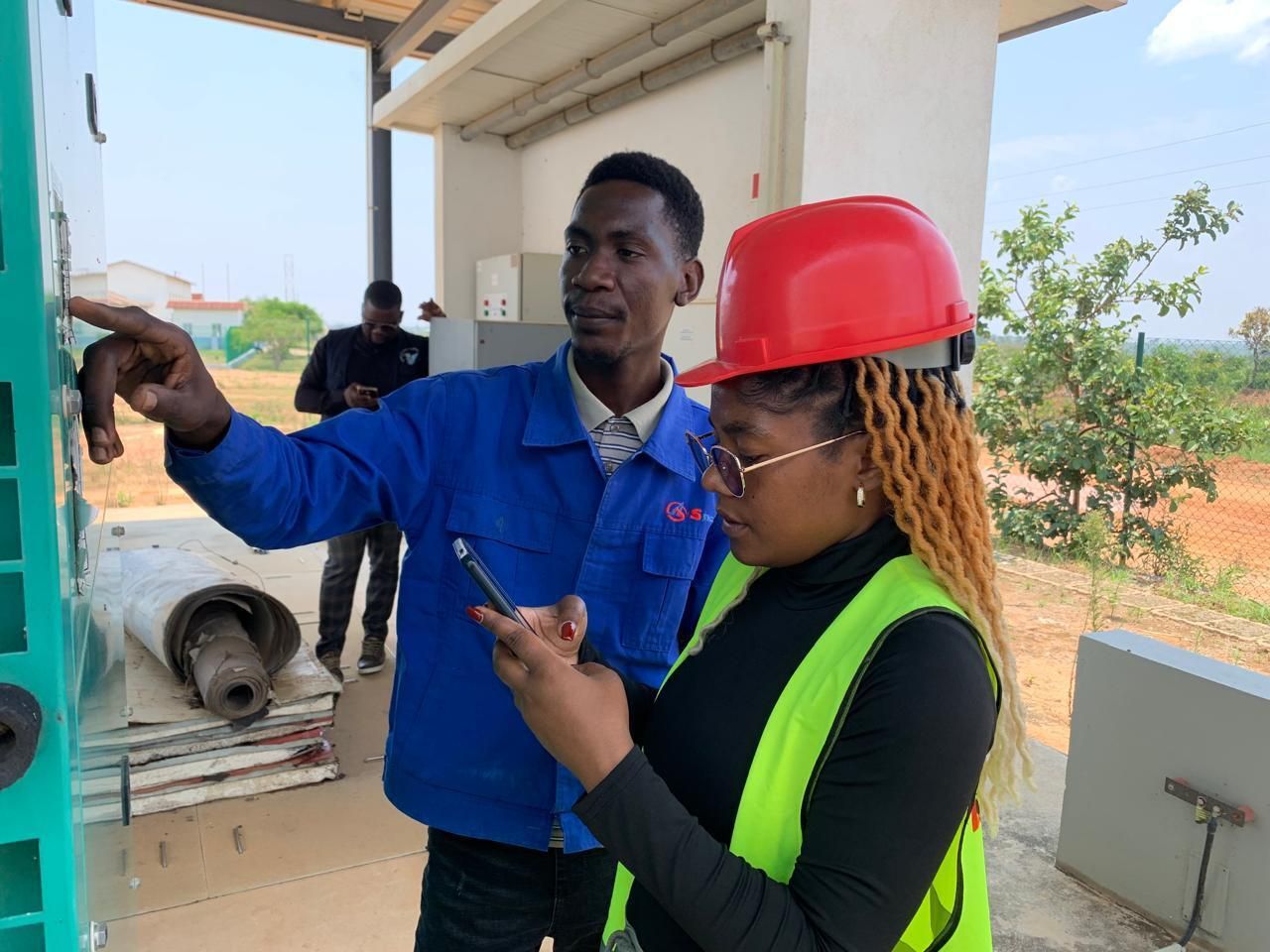 A utility technician shows an inspector the backup generator details to record on a phone, in Bié, Angola